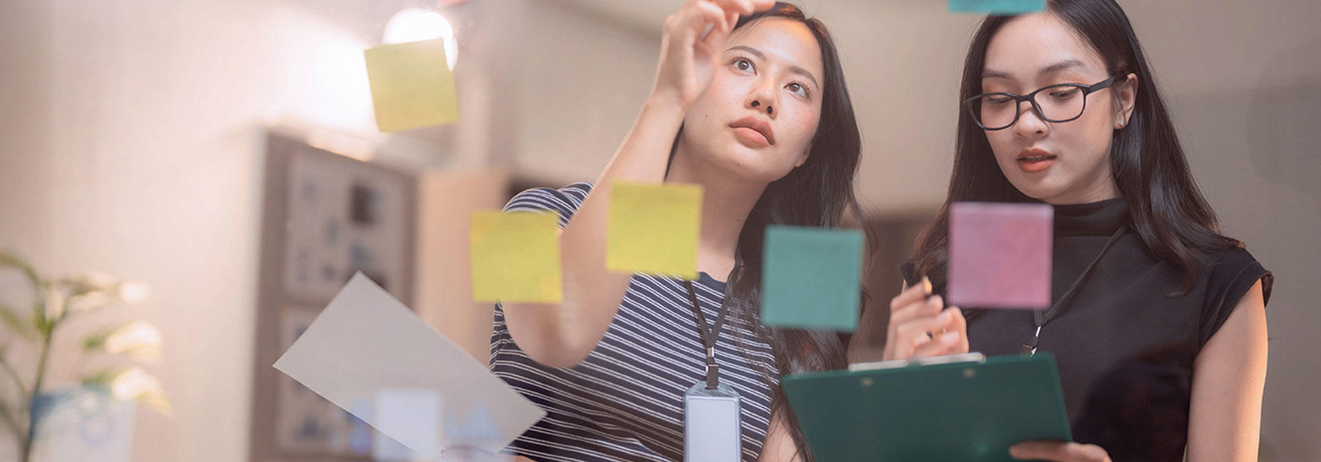Two women examine colorful sticky notes arranged on a wall, discussing ideas and collaborating on a project.