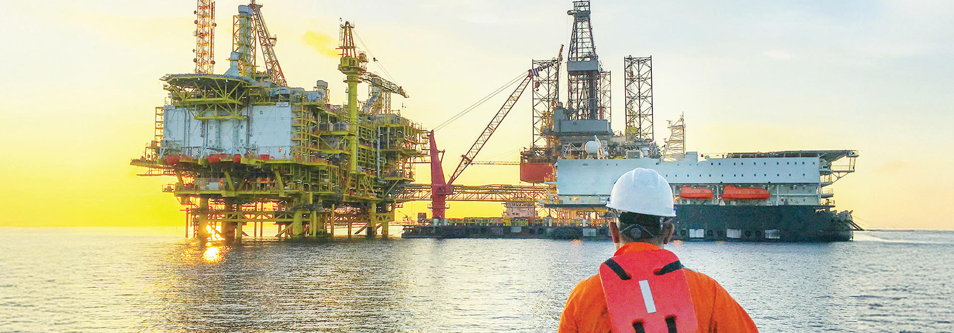 A man dressed in an orange safety vest stands before an oil rig, showcasing safety protocols in a construction setting.