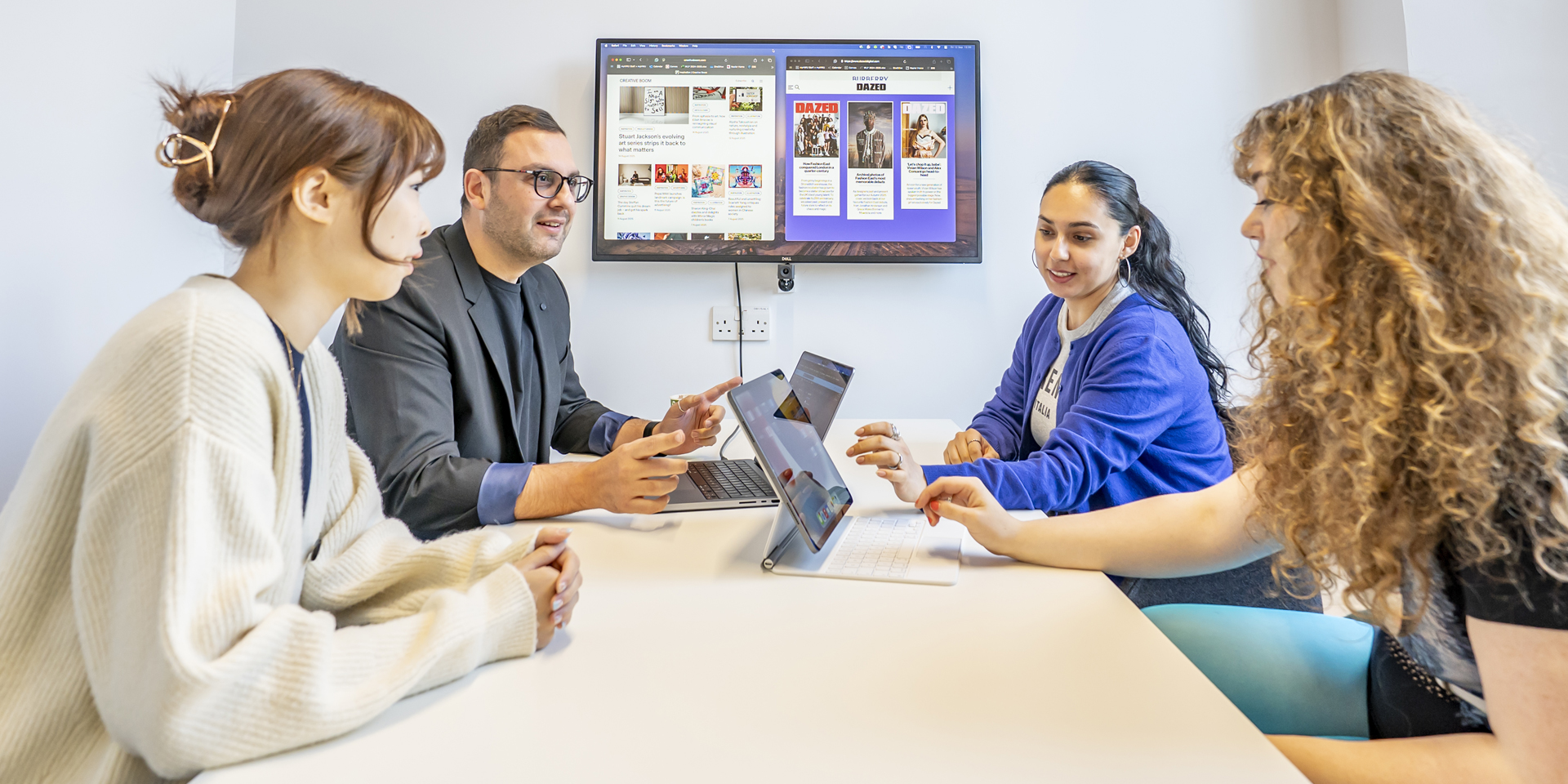 Four students at a table with laptops, focused on their work, while a television displays content in the background.