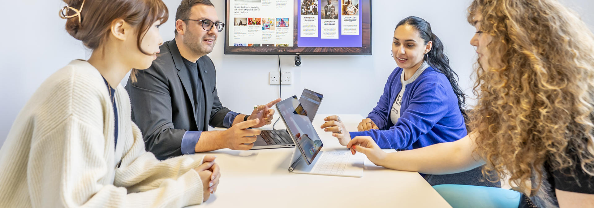 Four students at a table with laptops, focused on their work, while a television displays content in the background.