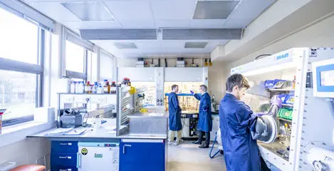 Two students in a chemistry lab, one using an isolation glove box and one talking with a member of staff