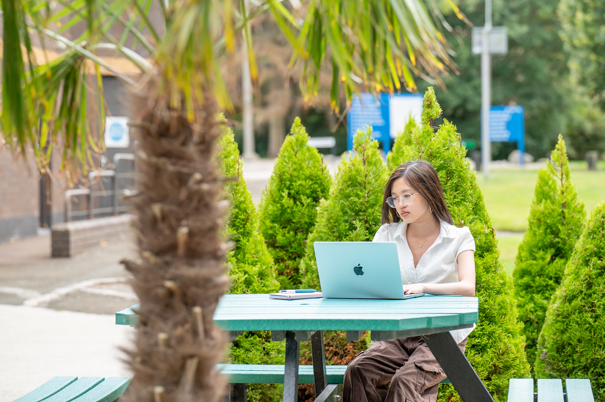 Female student sitting on a picnic table with laptop outside with greenery around