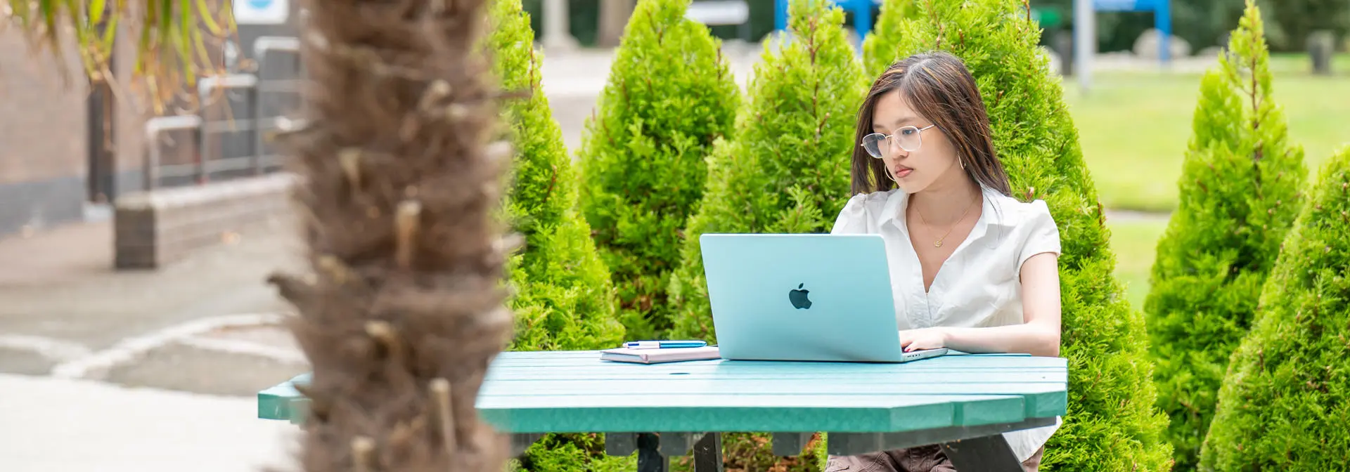 Female student sitting on a picnic table with laptop outside with greenery around
