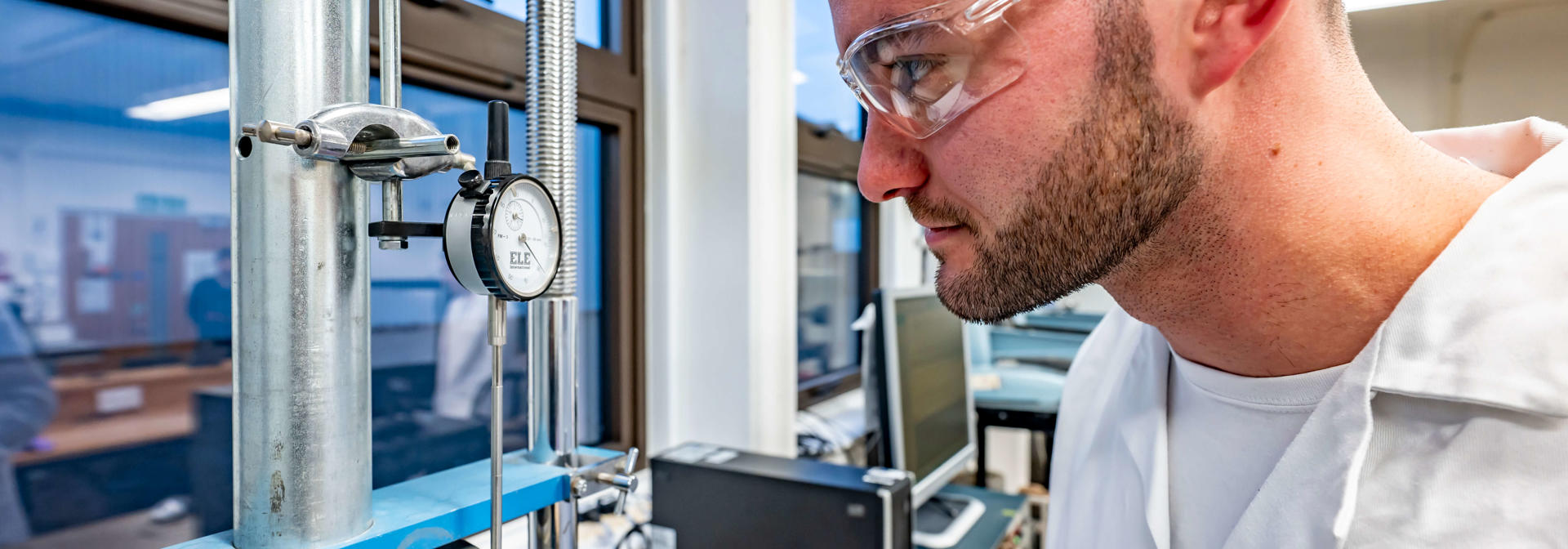 A student in white lab coat and safety glasses checks a pressure gauge