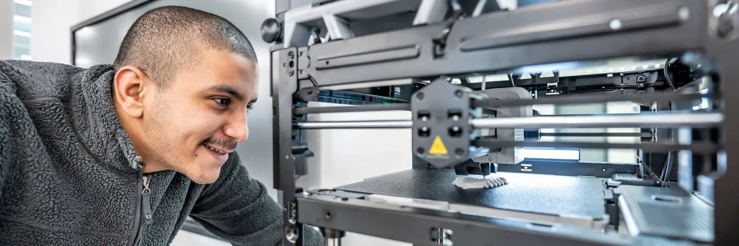A student smiling as he watches a 3D printed printing an object