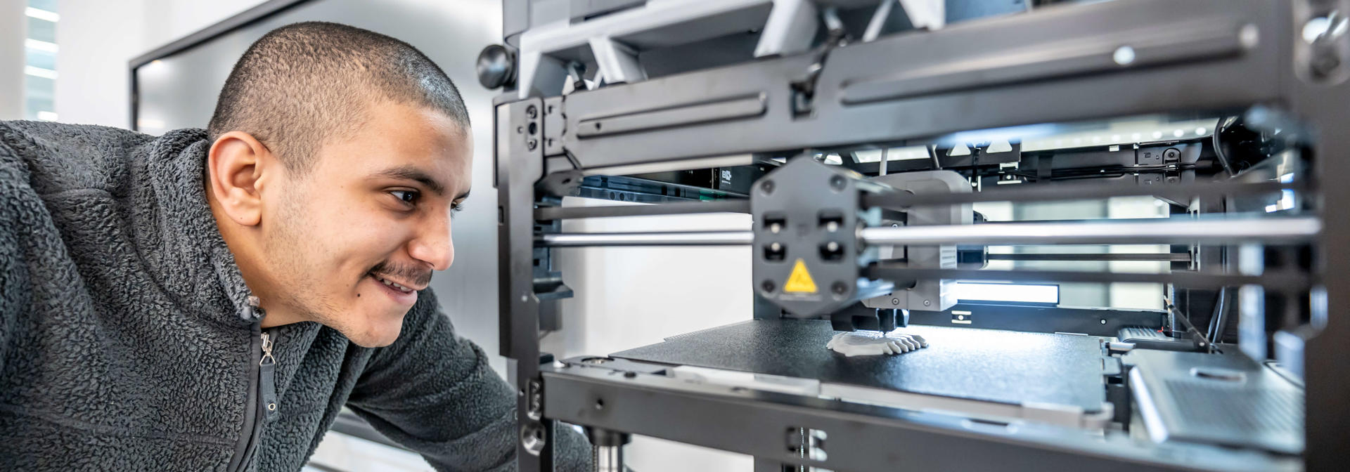 A student smiling as he watches a 3D printer printing an object