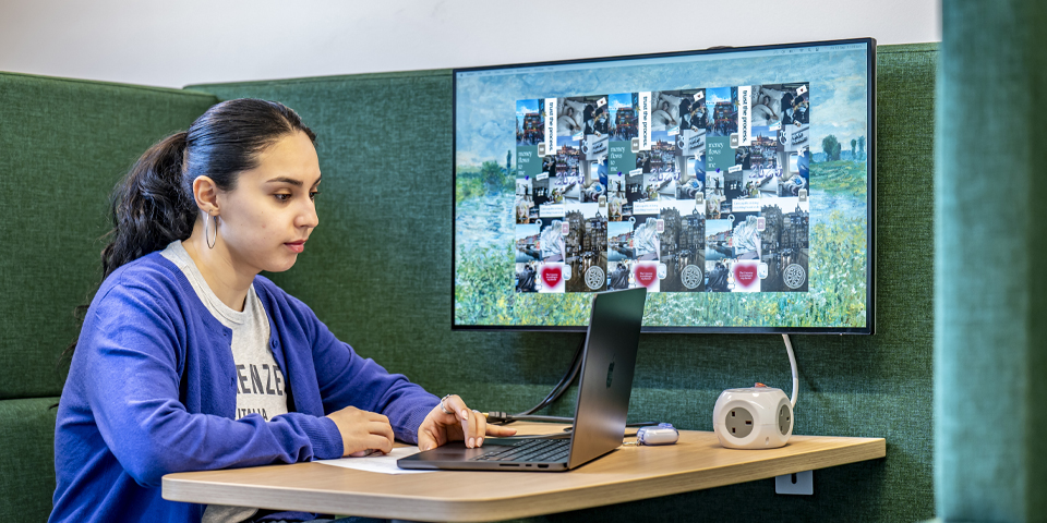 A student sits at a table with a laptop, focused on a monitor displaying a collage of images and graphics.