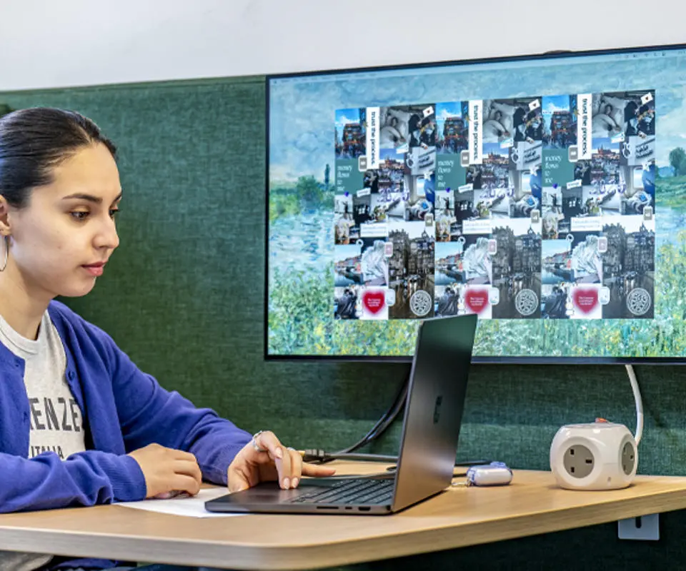 A student sits at a table with a laptop, focused on a monitor displaying a collage of images and graphics.