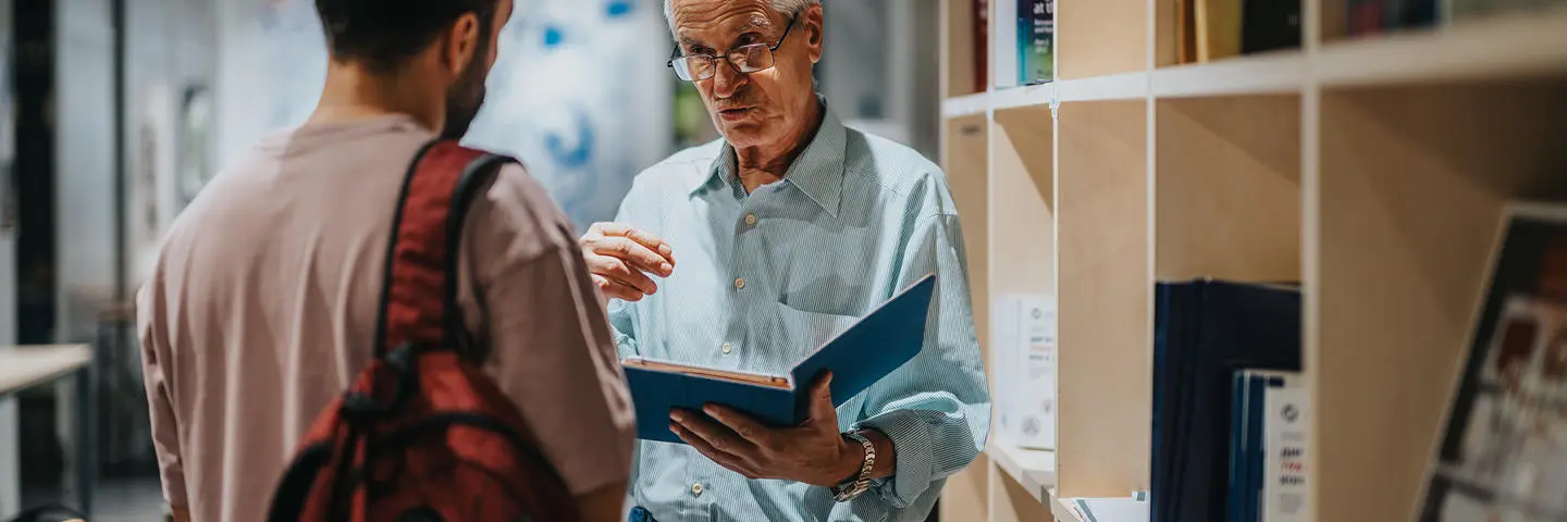 A coach talks through notebook guidance in the library to a student.