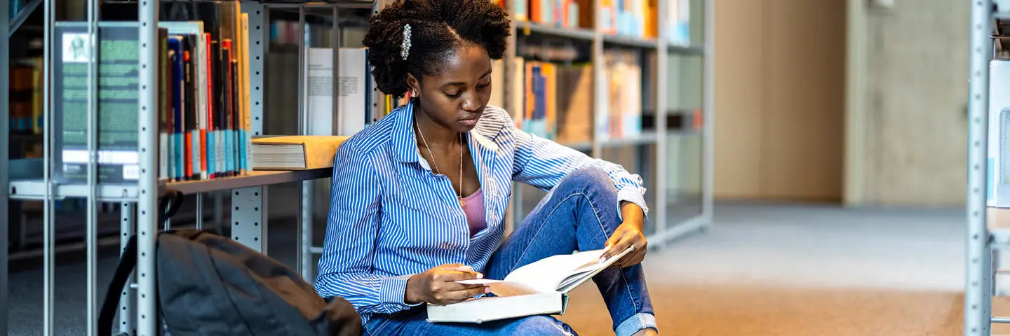 A student sits on the floor in the library reading a book.