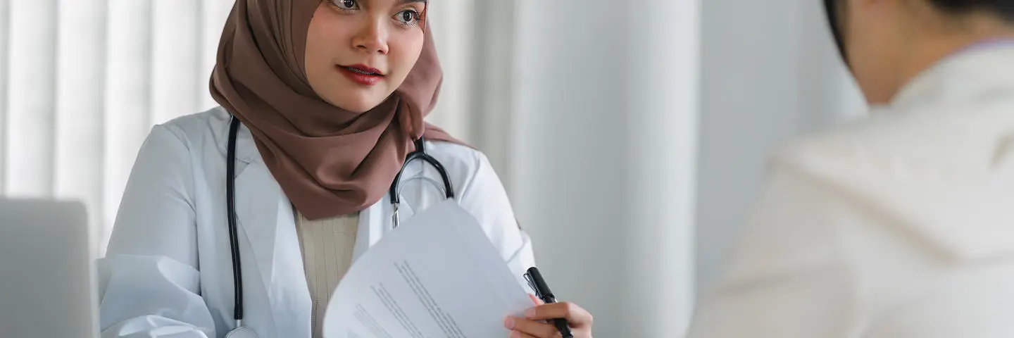 Doctor wearing a headscarf speaks at a desk to a person.