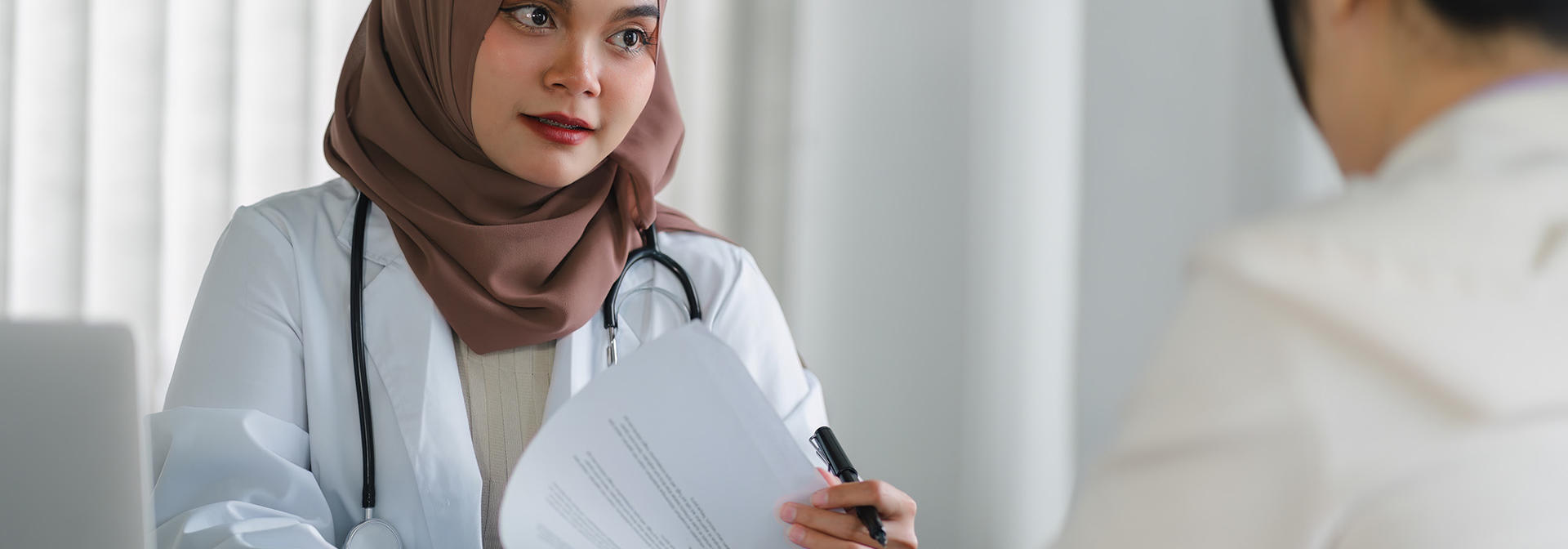 Doctor wearing a headscarf speaks at a desk to a person.