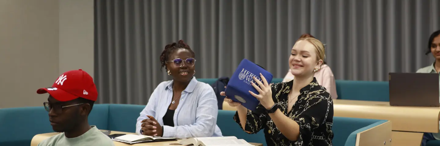 Three individuals sitting in a lecture theatre, smile as they pass around a cube.