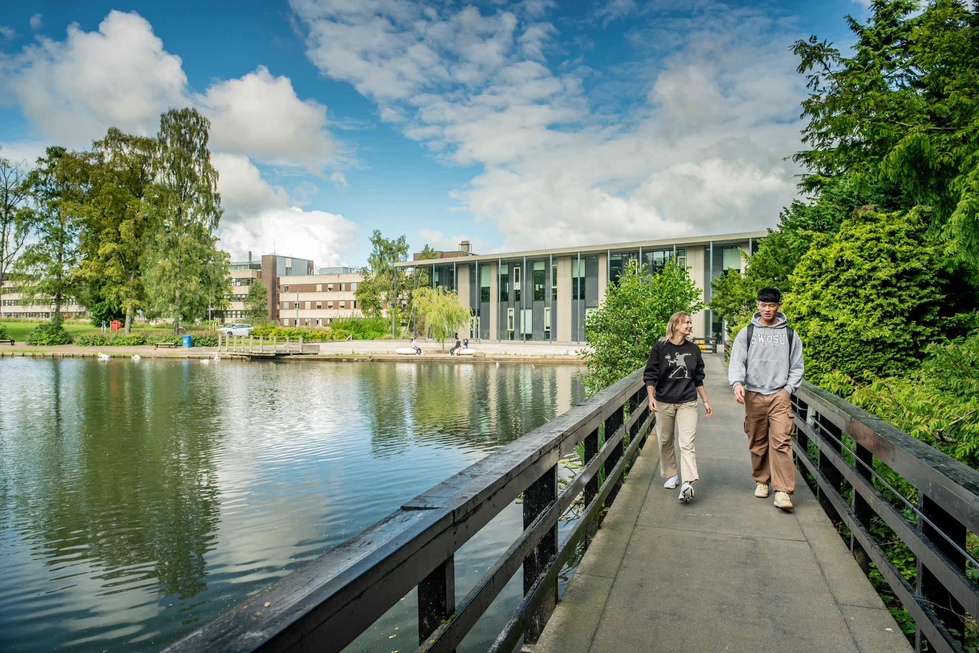 Tw individuals walking across a bridge above a loch on a sunny day.