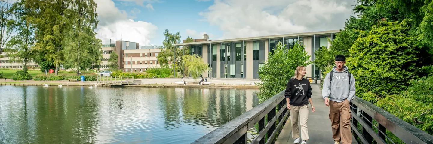 Tw individuals walking across a bridge above a loch on a sunny day.
