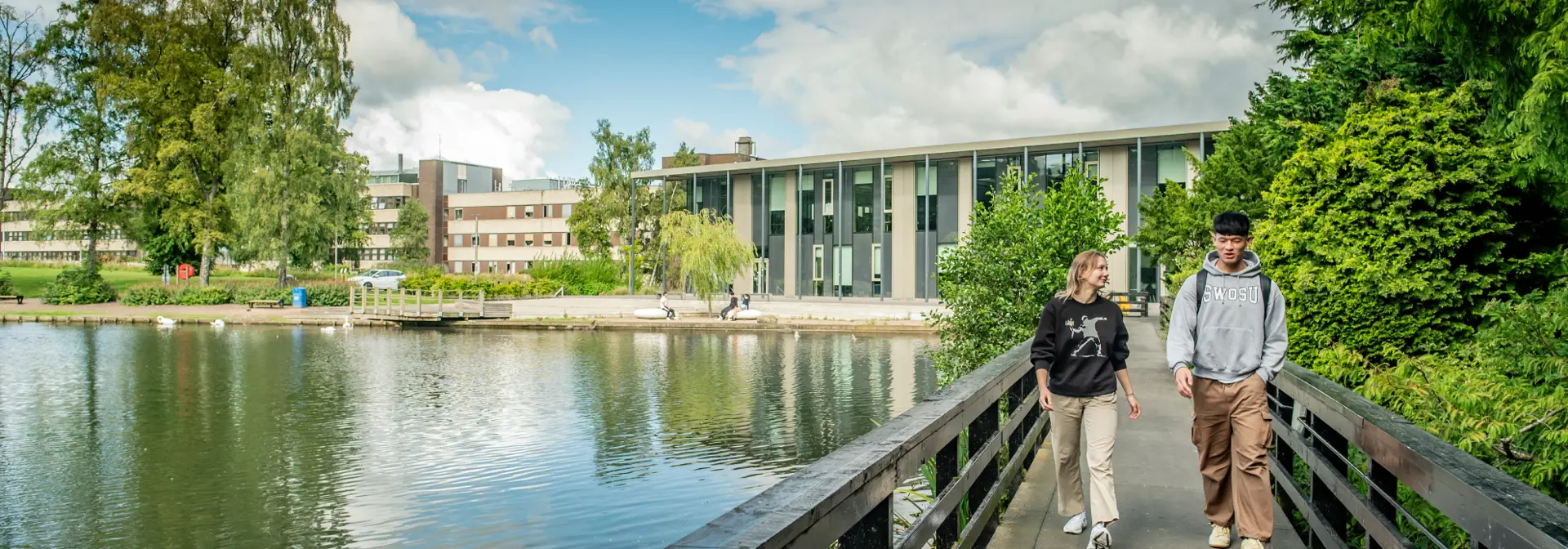 Tw individuals walking across a bridge above a loch on a sunny day.