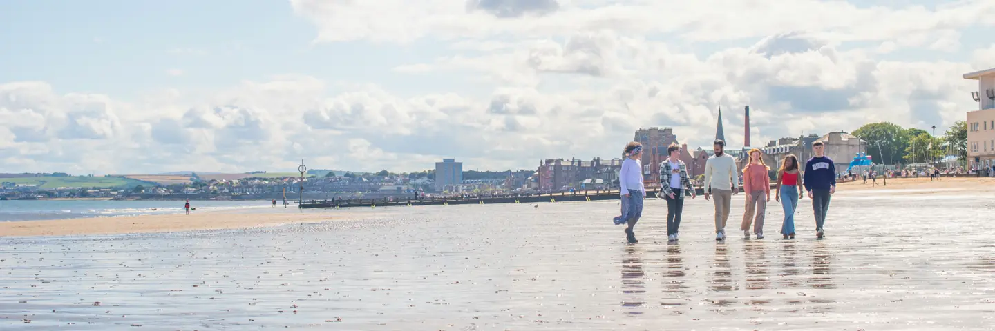 Five students walking on wet sand at Portobello beach with the town in the background.