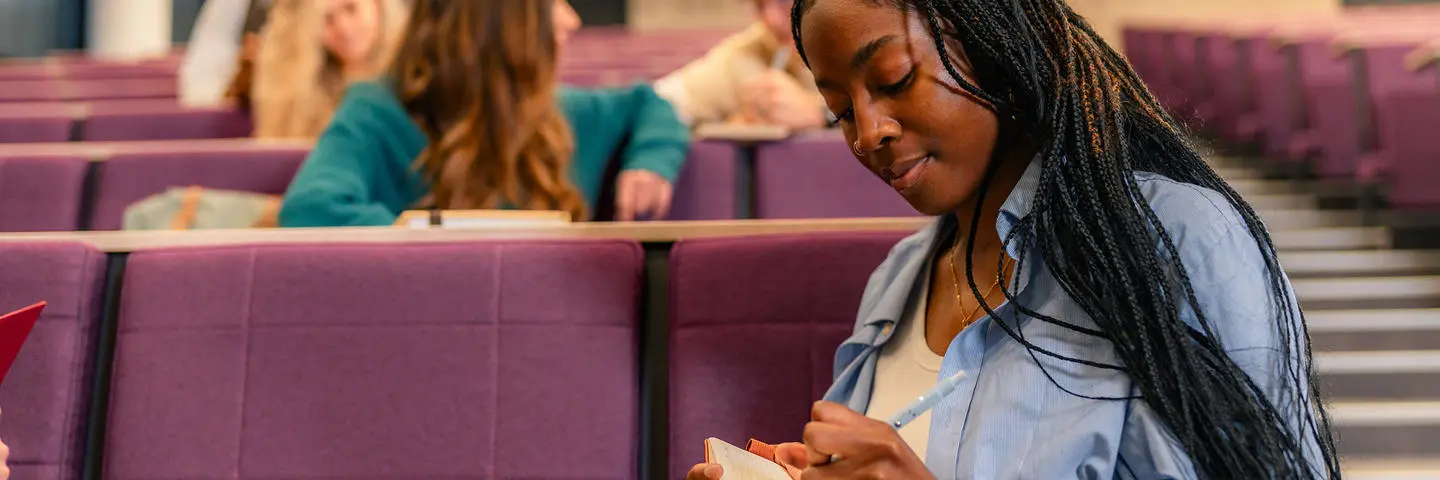 A student takes notes while sitting in a lecture hall.