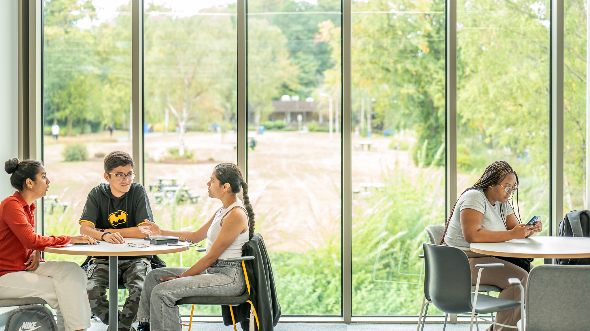 Students sitting at tables beside floor-to-ceiling windows which outlook onto the greenery on the Edinburgh campus.