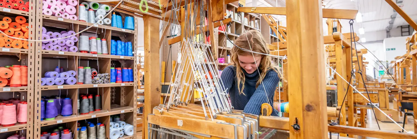 A woman works with threads and materials on a wooden weaving machine.