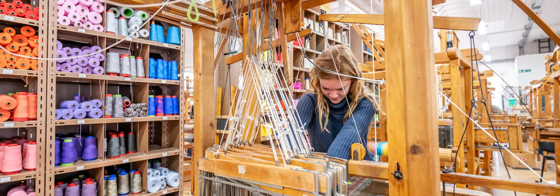 A woman works with threads and materials on a wooden weaving machine.