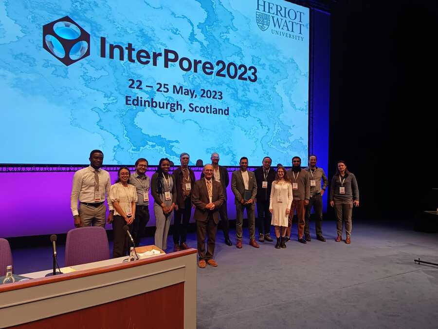 A group of 13 people stand together on a stage. Behind them is a holding slide promoting the InterPore 2023 Conference in Edinburgh from 22-25 May.