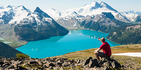 Lone male sitting on rocks on mountaintop looking at stunning scenery of snowclad mountains and a lake in the distance