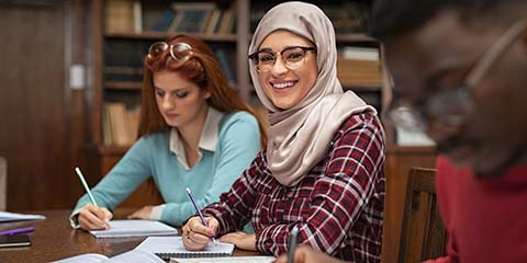 Smiling female student wearing abaya and spectacles while working in a university library.