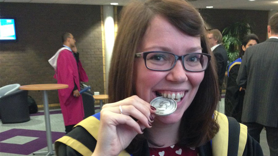 Medal recipient in graduation robes jokingly bites on the edge of her medal