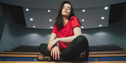 Female undergraduate student wearing flowery boots, black jeans and red t-shirt sits cross-legged on top of a lecture theatre desk