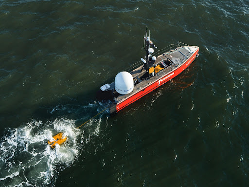 A cargo ship pulls an electric remote operated vehicle behind it at sea through choppy waters