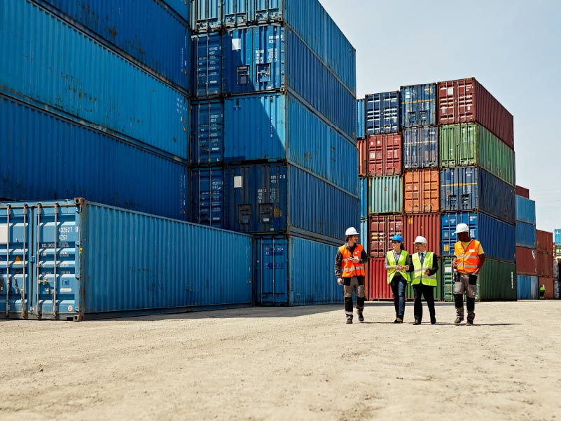 workers in high visibility jackets and helmets walking next to stacked shipping containers