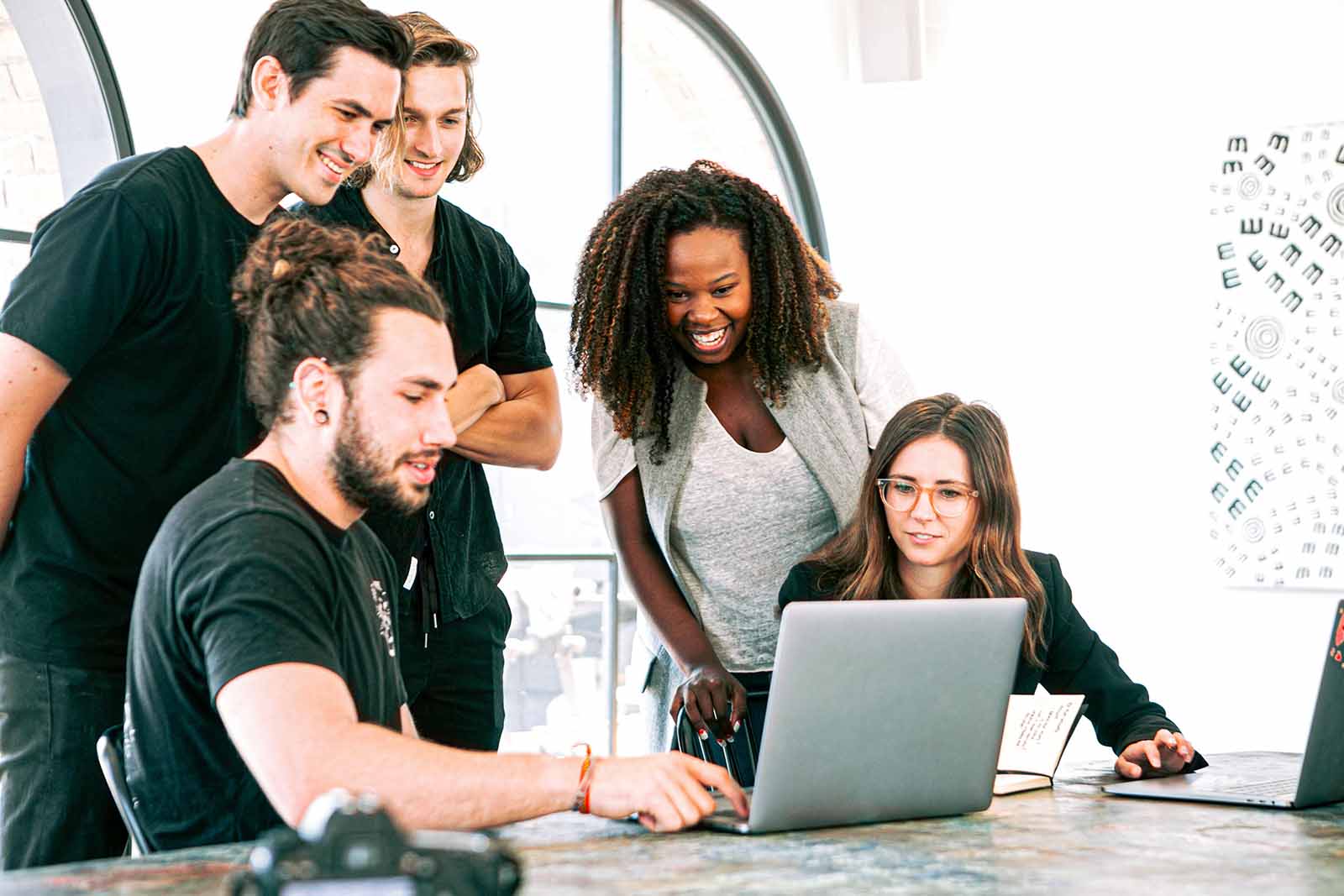 A group of people in an office huddles round a laptop smiling and laughing