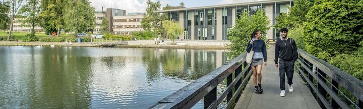 Two students strolling on a bridge above a loch, surrounded by lush greenery.