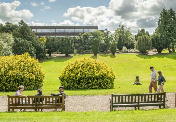 Individuals relax on benches by the lawn, framed by greenery from trees and bushes.