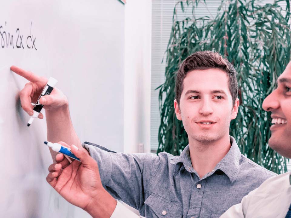 Two male colleagues discuss an equation while writing on a whiteboard