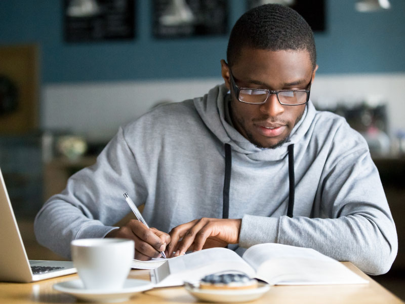Man wearing a hoodie and glasses takes notes from a textbook