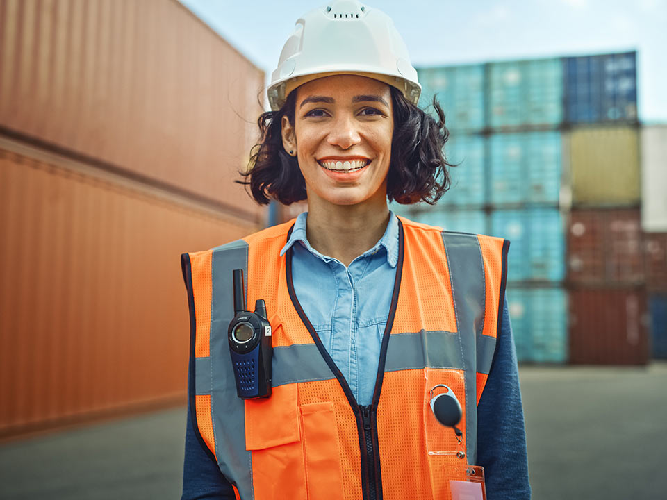 woman smiling at camera, wearing a construction helmet and high visibility vest, standing in front of shipping containers