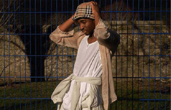 person standing against fence wearing beige draped clothing and a checked hat