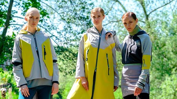Three female models standing at edge of river. Left model is wearing a grey waist length jacket with yellow hood and side panels. Middle model is wearing a full length yellow jacket with grey collar and sleeves. Right model is wearing a grey hoodie with a high roll neck and yellow arm band, with a grey mini skirt