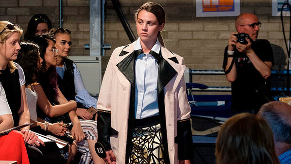 female model walking runway, wearing a white skirt, black and tan abstract triangle print mini skirt and an oversized black and tan blazer with a double collar