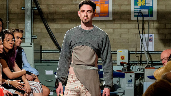 male model walking runway, wearing a cropped grey jumper over a brown tunic top with a silver belt
