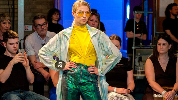 female model walking runway wearing, yellow ruched front top with green metallic high waist trousers and a full length light green jacket