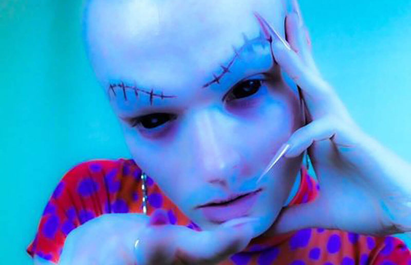 bald student with drawn-on stitched eyebrows, posing leaning on hand with long stiletto nails 