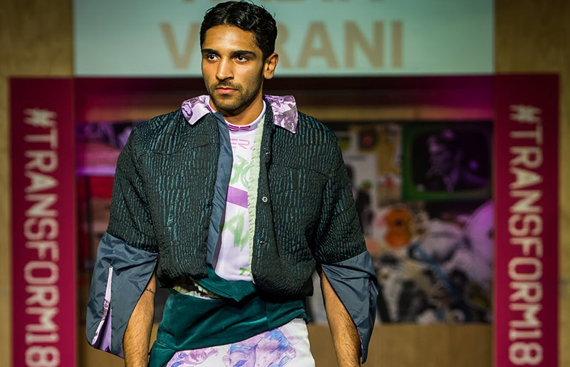male walking runway, wearing a purple and green printed top with a black and green striped jacket, which has cropped sleeves white flow from the elbow in a black satin