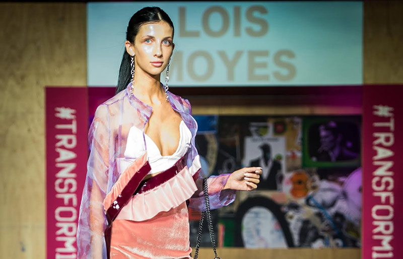 female walking catwalk, wearinga purple iridescent jacket with pink and red frills, a white bralette and a light pink velvet mini skirt 