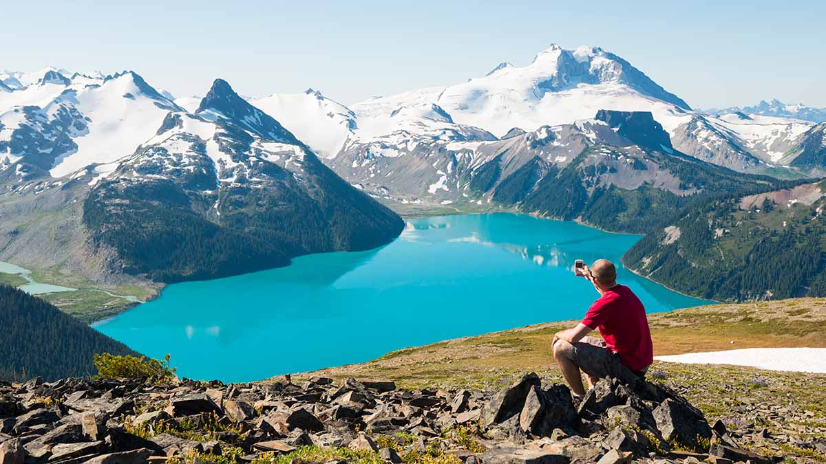 Lone male sitting on rocks on mountaintop looking at stunning scenery of snowclad mountains and a lake in the distance