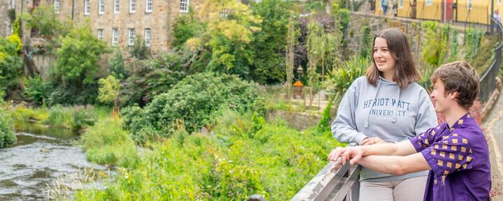Two young students stand, gazing at the flowing river below, surrounded by a serene natural landscape of Edinburgh's Dean Village